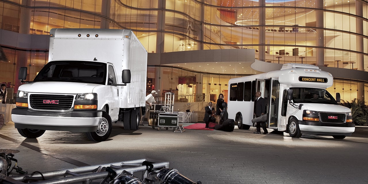 Two GMC Commercial Savana Cutaway Vans Parked in Front of a Large Lite up Building at Night