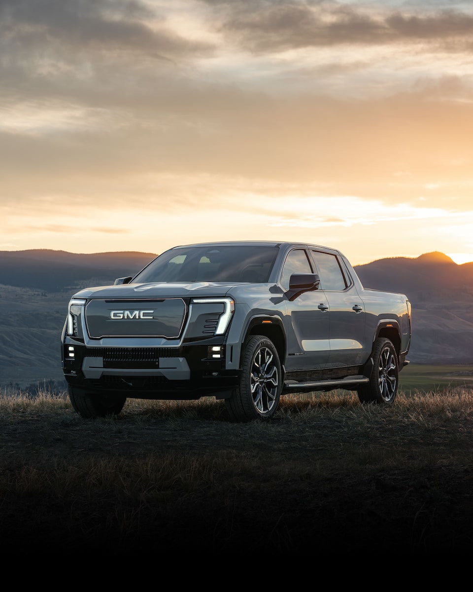 Front Three-Quarters View of a GMC Sierra Denali Edition 1 Electric Truck Driving on an Open Road with Mountains in the Background