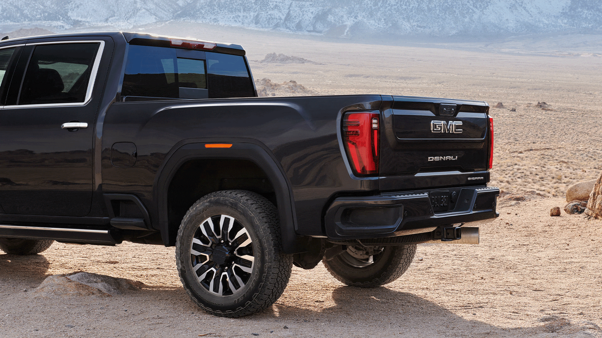 Rear Three-Quarter View of a Dark Gray GMC Sierra Truck with its Tailgate Lowered Parked on a Dirt Path with Rugged Tires and Mountains in the Background