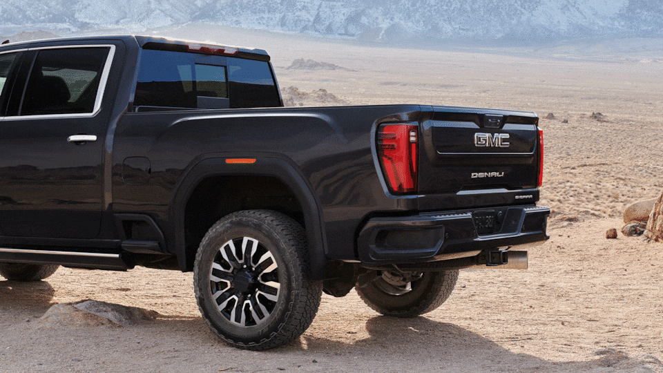 Rear Three-Quarter View of a Dark Gray GMC Sierra Truck with its Tailgate Lowered Parked on a Dirt Path with Rugged Tires and Mountains in the Background