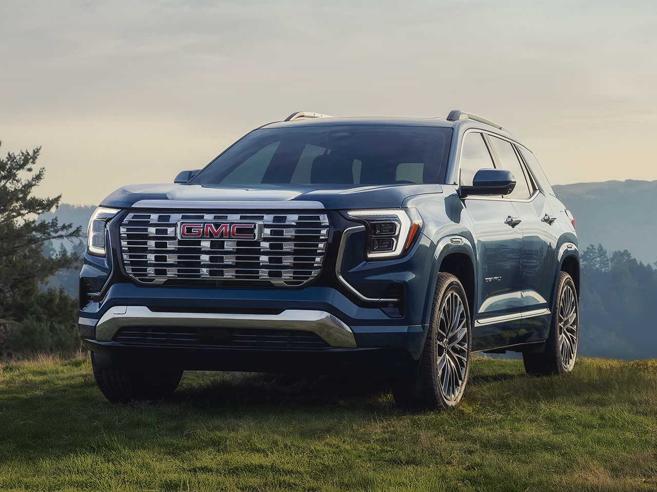Front Three-quarters View of a Dark Blue GMC Terrain SUV Parked on a Grassy Hillside with Mountains and Pine Trees in the Distance