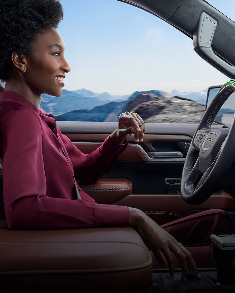 A Woman Smiling While Driving a GMC Truck with Hands Off the Steering Wheel, Viewed From the Passenger Side, with Mountains in the Background, and an Interior Featuring Brown Leather and a Large Screen