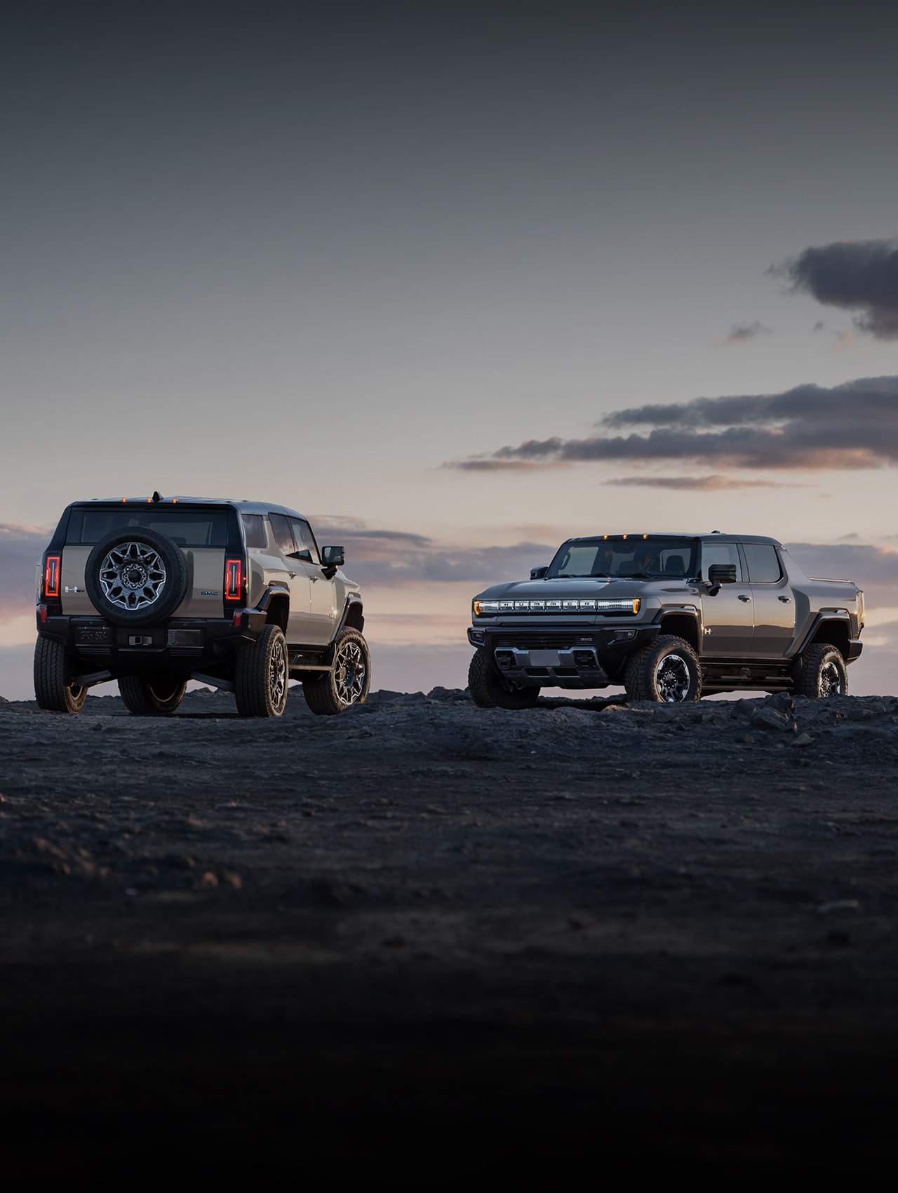 The GMC HUMMER EV SUV and the GMC HUMMER EV Pickup Truck Parked on Rough Terrain Over Looking a Beautiful Sunset Colored Sky