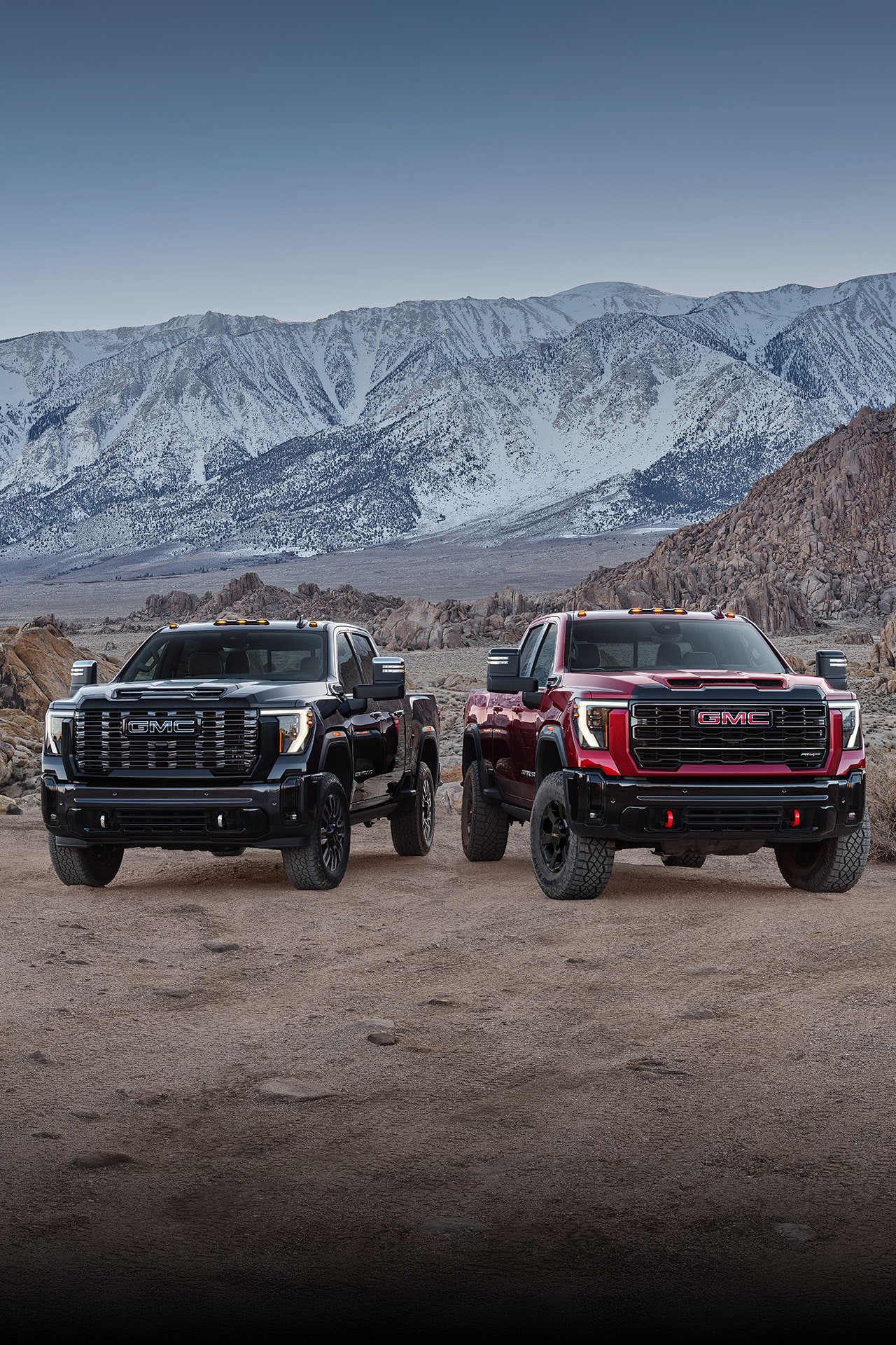 Two 2027 GMC Sierra HD Trucks, One Black and One Red, with Off-Road Tires and Red Recovery Hooks Parked Side By Side On a Rocky Terrain with Snowy Mountains in the Background