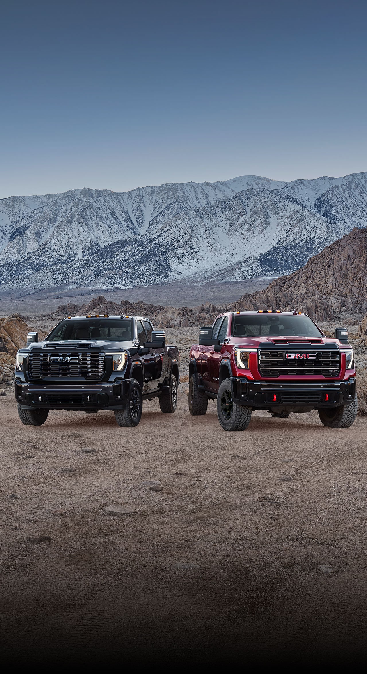 Two 2027 GMC Sierra HD Trucks, One Black and One Red, with Off-Road Tires and Red Recovery Hooks Parked Side By Side On a Rocky Terrain with Snowy Mountains in the Background