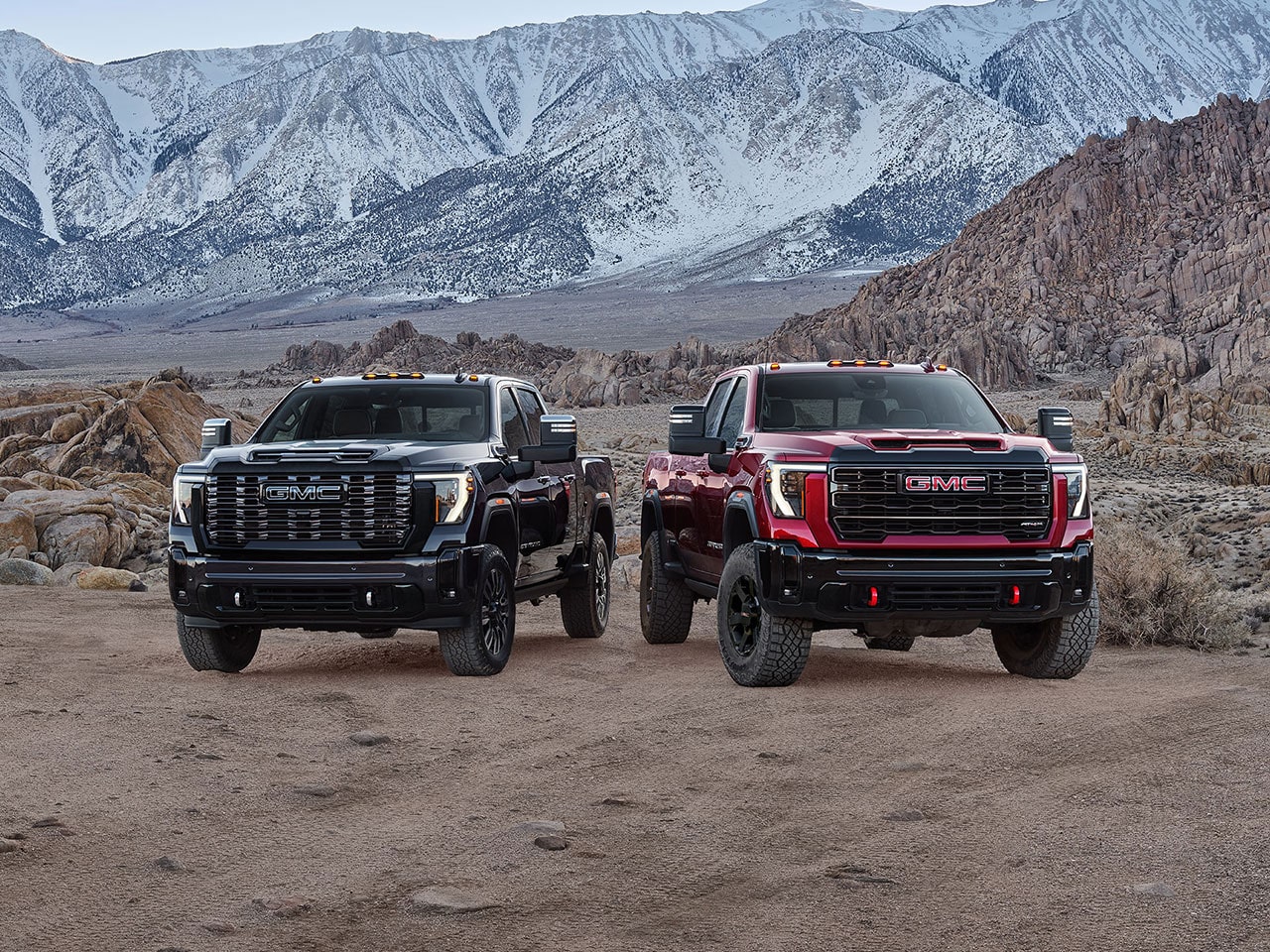 Two 2027 GMC Sierra HD Trucks, One Black and One Red, with Off-Road Tires and Red Recovery Hooks Parked Side By Side On a Rocky Terrain with Snowy Mountains in the Background