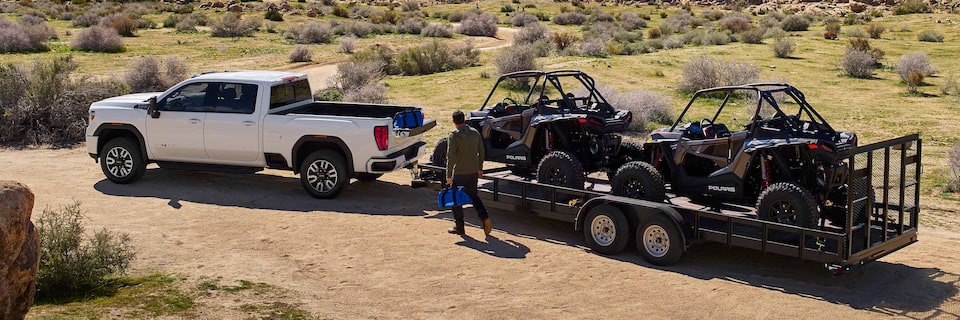 Profile View of a GMC Sierra HD Parked on a Dirt Road Towing a Trailer with Two Polaris Off-Road Vehicles