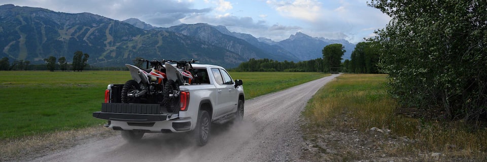 Rear View of a GMC Sierra AT4 Truck Driving Down a Dirt Road with Two Dirt Bikes in the Truck Bed