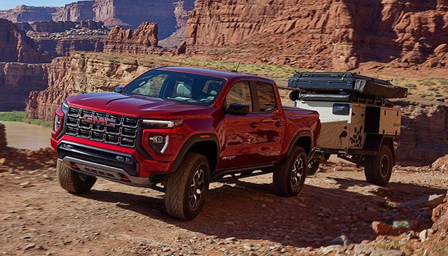 A Red GMC Canyon Pickup Truck Towing an Adventure Trailer Over a Rocky, Uneven Dirt Road in a Canyon