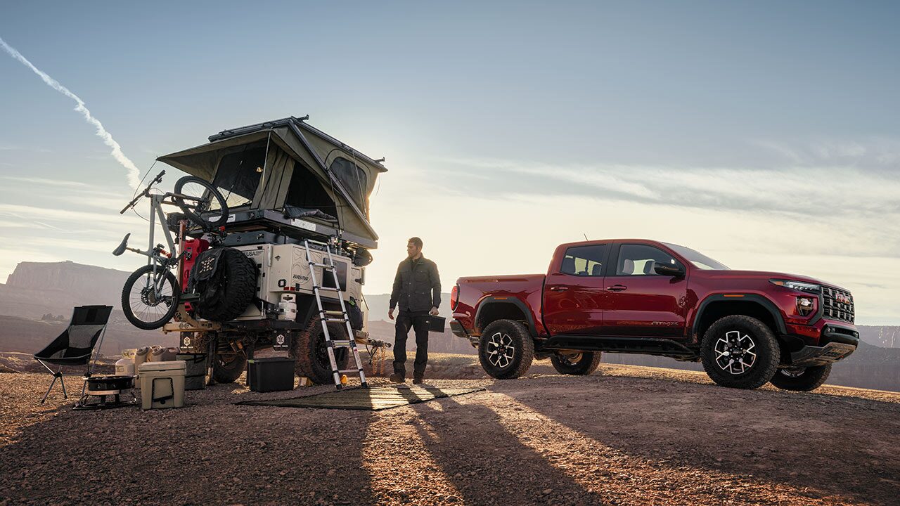 A Red GMC Canyon Pickup Truck and a Man Standing Next to an Adventure Trailer with a Rooftop Tent and Bicycle in a Desert Landscape