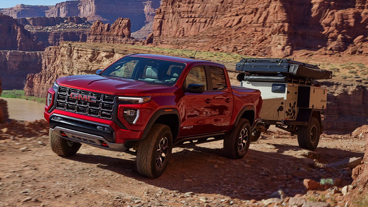 A Red GMC Canyon Pickup Truck Towing an Adventure Trailer Over a Rocky, Uneven Dirt Road in a Canyon
