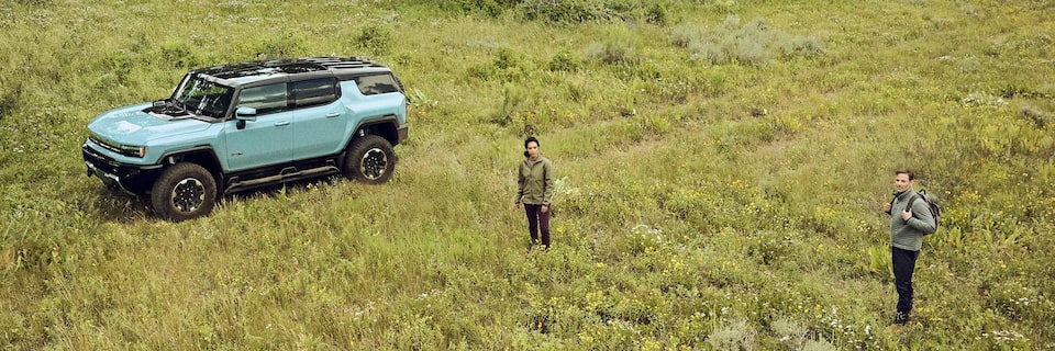 A Couple in Hiking Gear Standing in a Green Meadow with Their HUMMER EV Parked Next to Them