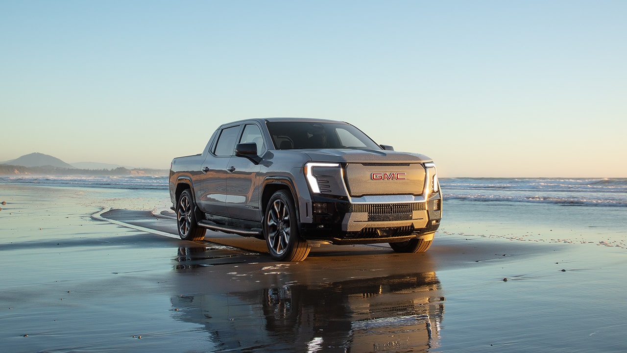 A Gray GMC Sierra EV Pickup Truck Parked on a Wet Sandy Beach at Sunset with the Ocean in the Background