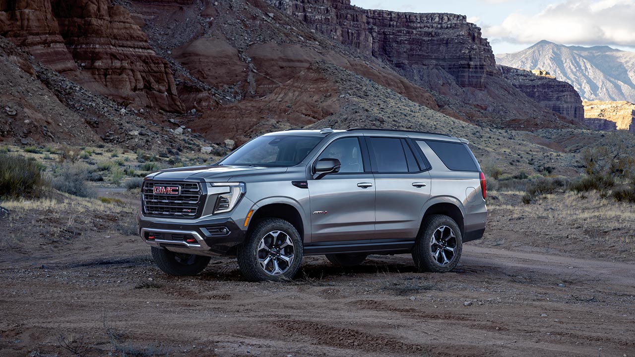A Light Gray GMC Yukon AT4 SUV Is Parked on a Dirt Trail in a Rugged Desert Landscape With Cliffs and Sparse Vegetation Under an Overcast Sky