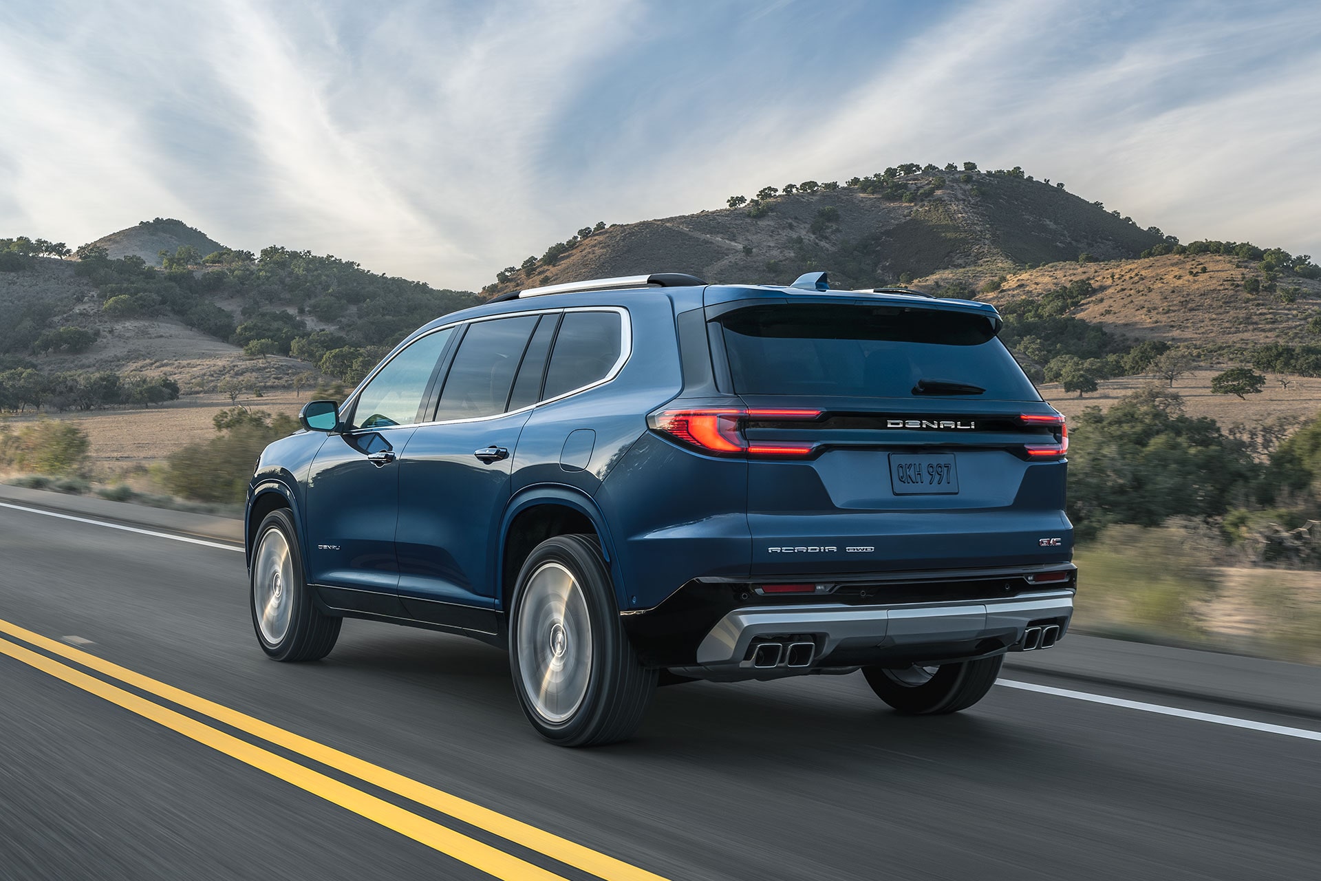 The Rear View of a Blue GMC Acadia Denali Driving on a Two-Lane Road with Mountains in the Background