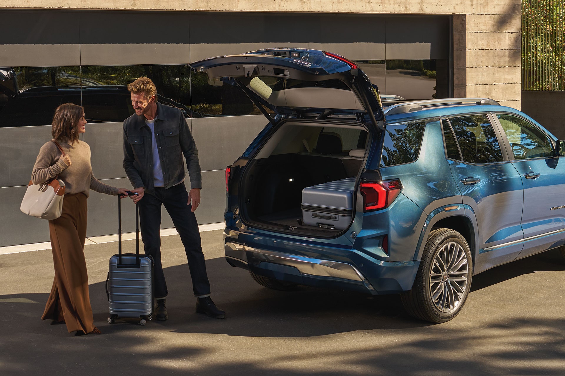 A Man and a Woman Loading Luggage into the Open Trunk of a Blue GMC Acadia SUV Parked in a Driveway