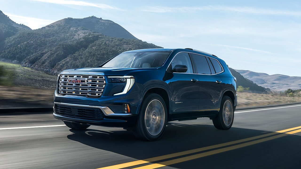 A Blue GMC Acadia SUV Driving on a Highway with Rolling Hills and a Clear Sky in the Background