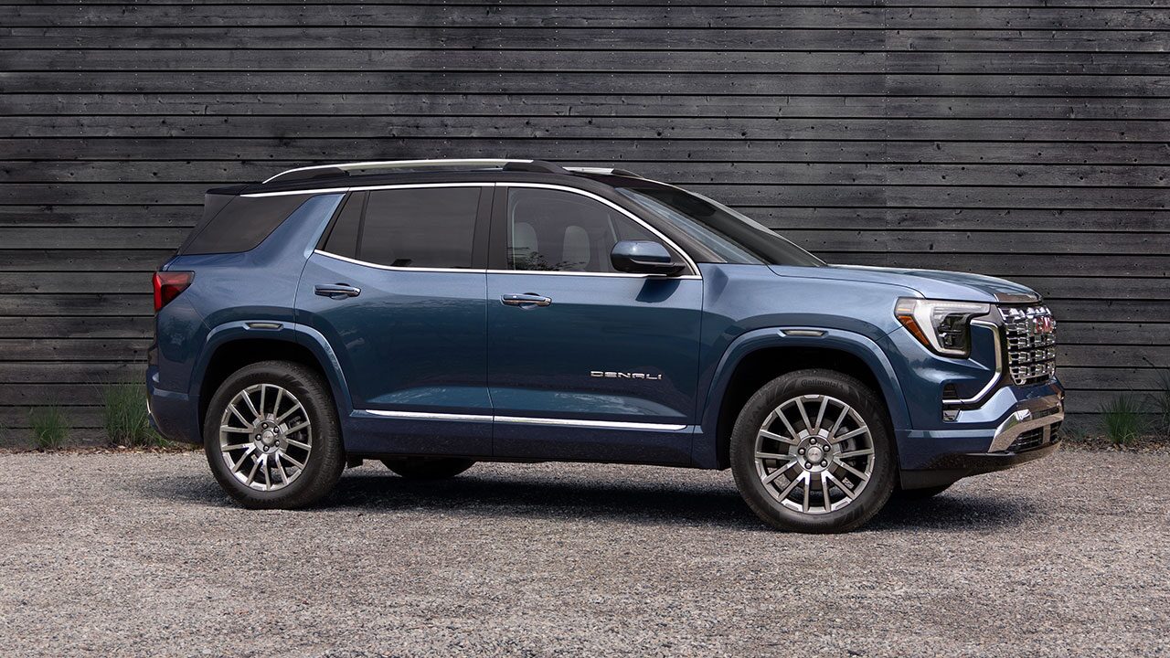 A Side Profile of a Blue GMC Acadia Denali SUV Parked on a Gravel Lot in Front of a Dark Wooden Wall