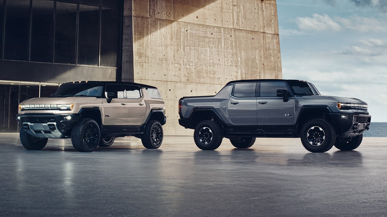 A Tan GMC Hummer EV SUV Parked Next to a Gray GMC Hummer EV Pickup Truck by a Modern Building and the Ocean