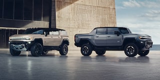 A Tan GMC Hummer EV SUV Parked Next to a Gray GMC Hummer EV Pickup Truck by a Modern Building and the Ocean