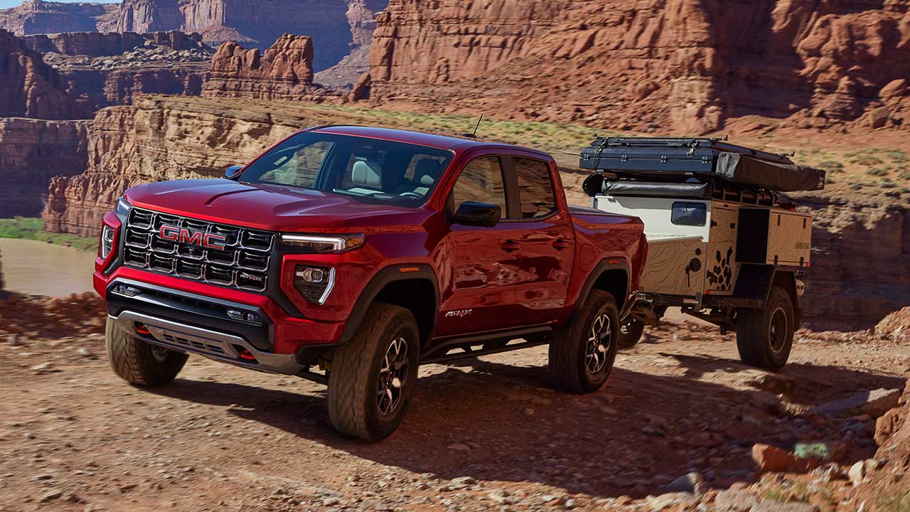 A Dark GMC Sierra HD AT4X AEV Edition Truck Is Kicking Up Dust While Driving on a Dirt Trail Through a Field of Dry Grass, With Mountains in the Background