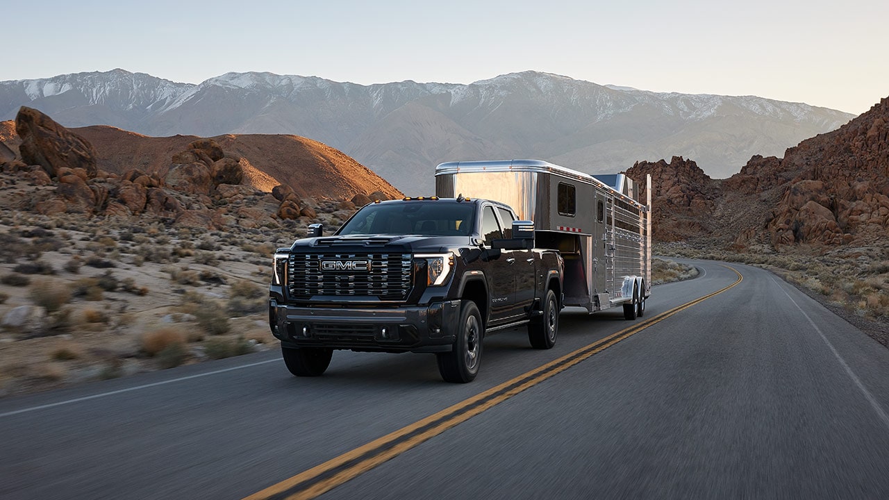 A Dark GMC Sierra Truck Is Towing a Large, Silver Horse Trailer Along a Winding Paved Road Through a Rugged Desert Landscape With Mountains
