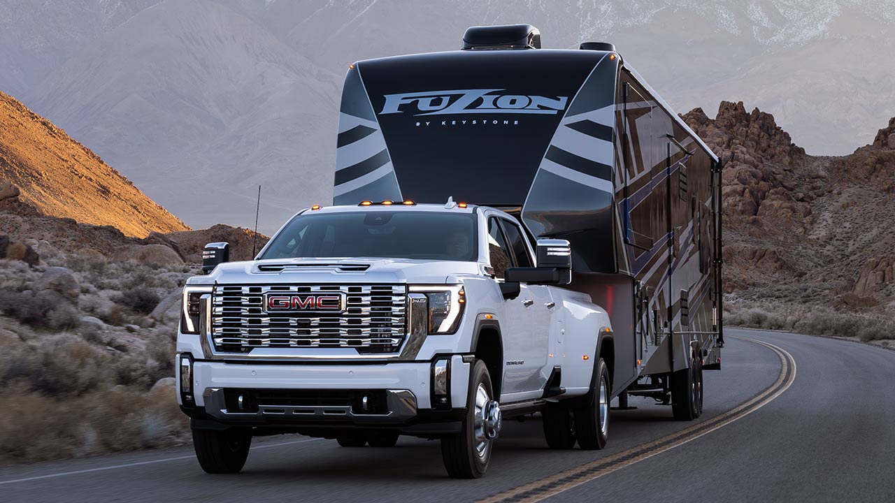 A White GMC Sierra Heavy-Duty Truck With a Large Fuzion by Keystone Fifth-Wheel Trailer Is Driving on a Paved Road Through a Rugged, Mountainous Desert Environment