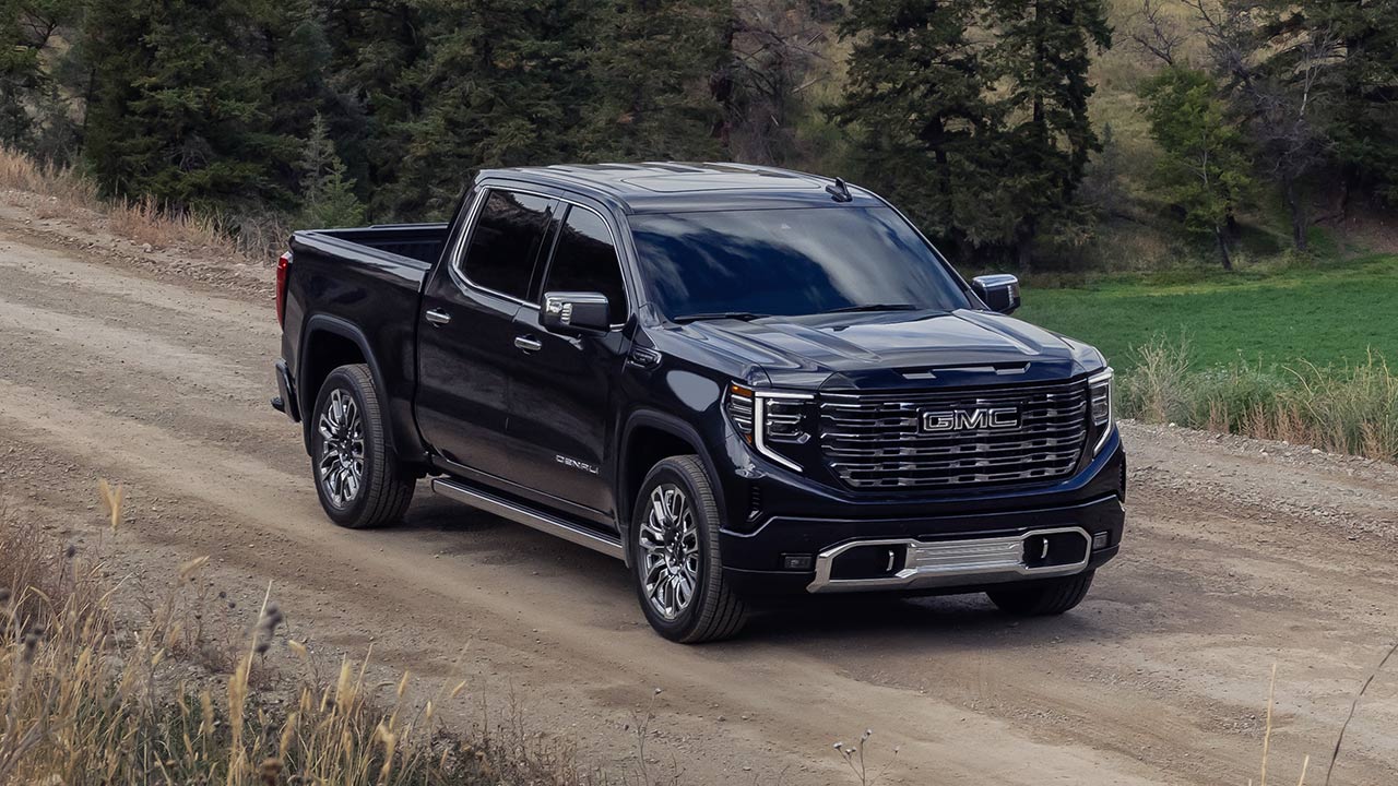 A Dark GMC Sierra Denali Truck Is Driving on a Dirt Road Through a Rural Landscape With Green Fields and Trees on Rolling Hills