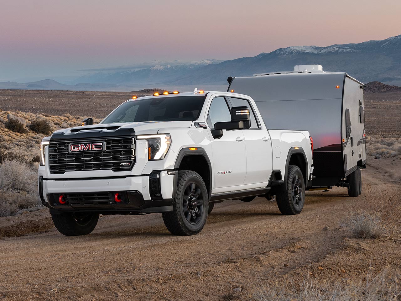 A White GMC Sierra AT4X Heavy-Duty Truck Is Pulling a Compact Travel Trailer on a Dirt Road in a Desert Landscape With Mountains in the Background