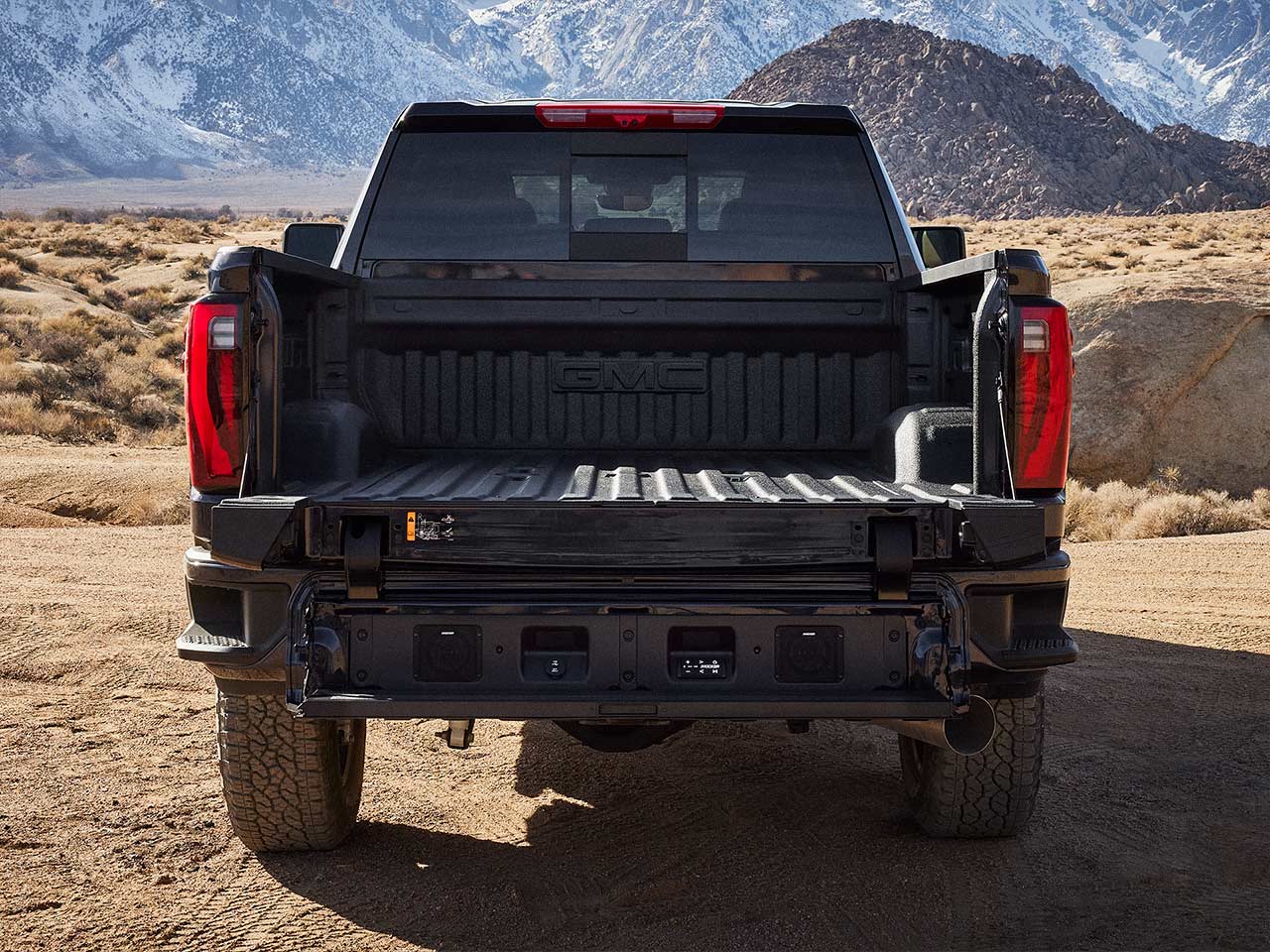 The Rear View of a Dark GMC Sierra Truck With Its Multi-Functional Tailgate Fully Opened, Revealing a Flat Cargo Bed and Integrated Steps, Parked in a Desert Landscape