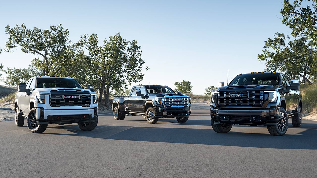 Three New GMC Sierra Heavy-Duty Trucks, One White and Two Dark, Are Parked Side-by-Side on a Paved Road With Sand Dunes and Sparse Vegetation in the Background