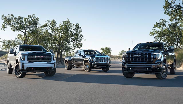 Three New GMC Sierra Heavy-Duty Trucks, One White and Two Dark, Are Parked Side-by-Side on a Paved Road With Sand Dunes and Sparse Vegetation in the Background