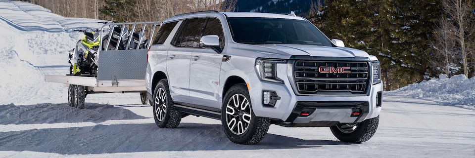 Three-Quarters View of a White GMC SUV Pulling a Trailer with Snowmobiles on a Road Covered by Snow