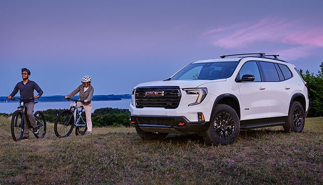 A White GMC SUV Parked on a Grassy Hill with a Man and a Woman Standing with Bicycles by a Lake at Sunset