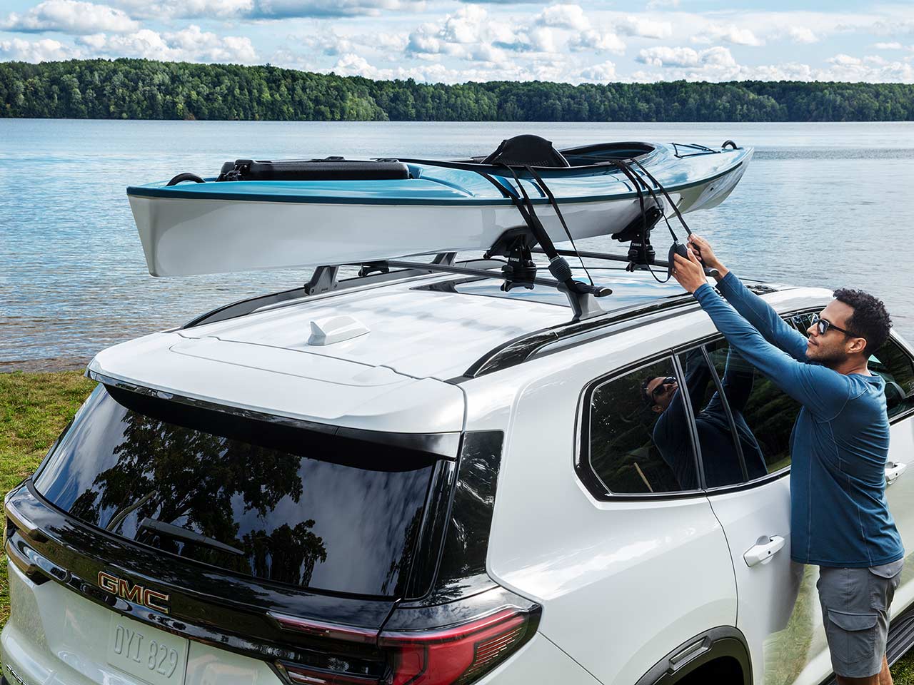 A Man Securing a Light Blue and White Kayak onto the Roof Rack of a White GMC SUV by a Lake