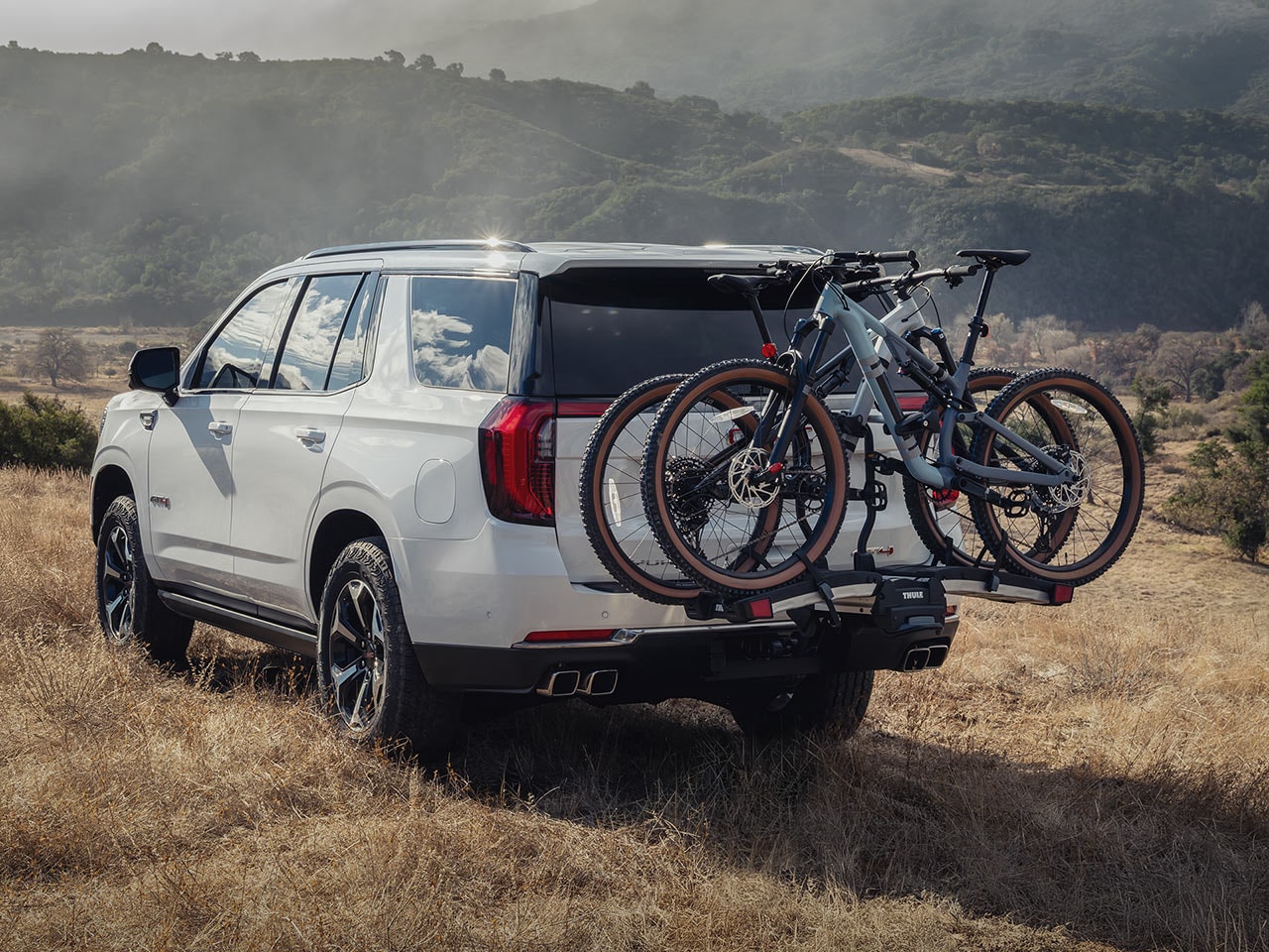 A White GMC Yukon SUV with a Bike Rack Carrying Two Bicycles in a Dry and Grassy Landscape