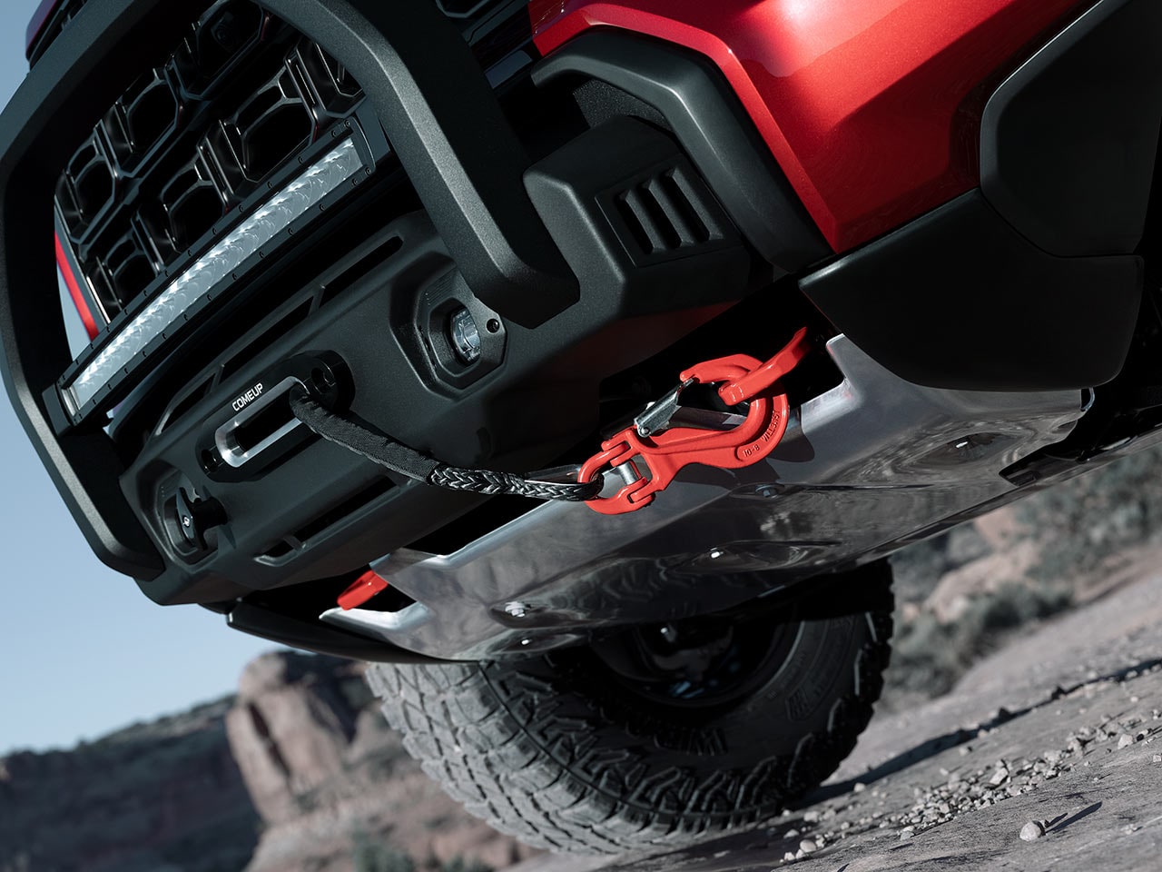 A Low-Angle View of a Red GMC Truck's Undercarriage Showing a COMEUP Winch and a Red Tow Hook on Rugged Terrain