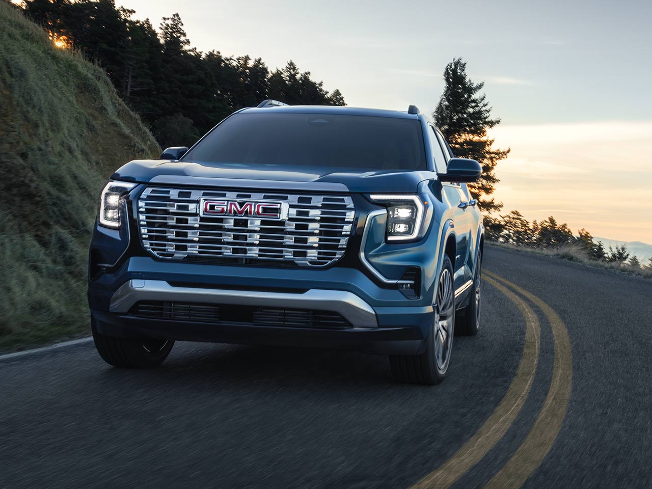 A Blue GMC SUV Driving on a Winding Road with Trees and a Sunset in the Background