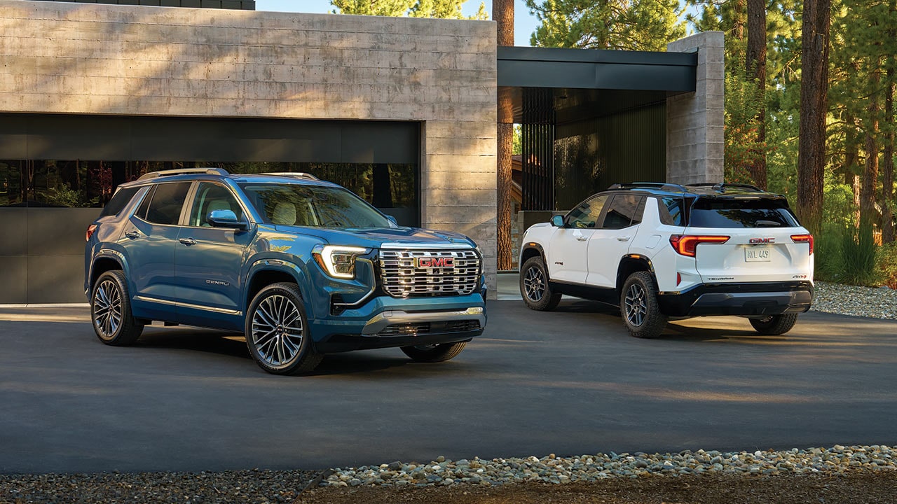 Two GMC SUVs Parked on a Paved Driveway in Front of a Modern House a Blue GMC Denali in the Foreground and a White GMC Terrain AT4 in the Background