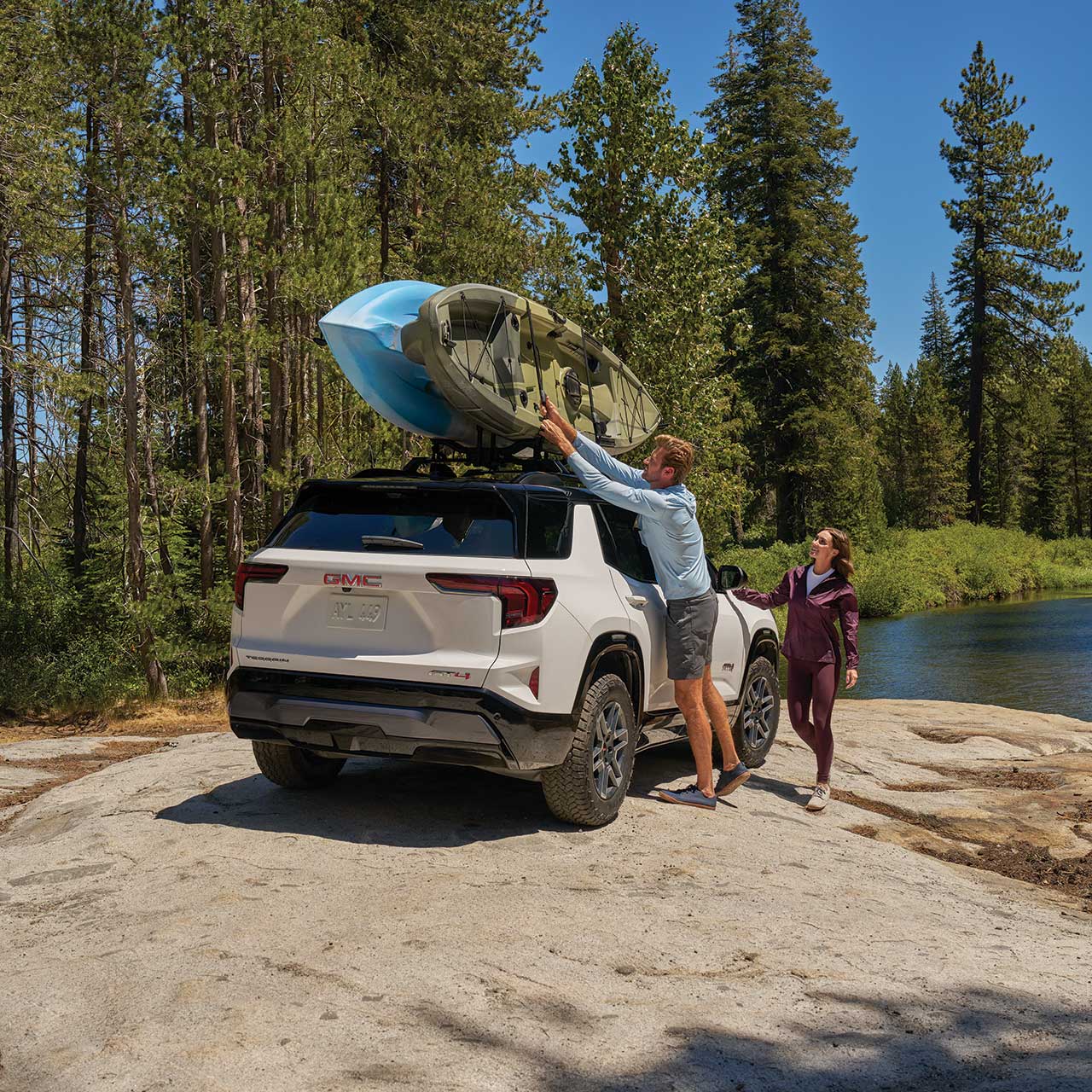 A White GMC Terrain SUV Parked by a Lake with Two People Putting a Kayak on the Roof Rack Surrounded by Tall Pine Trees
