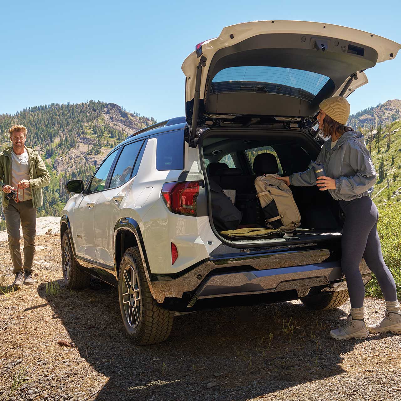 A White GMC Terrain AT4 SUV with Its Trunk Open a Woman Loading Gear Inside while a Man Stands Nearby in a Mountainous Outdoor Setting