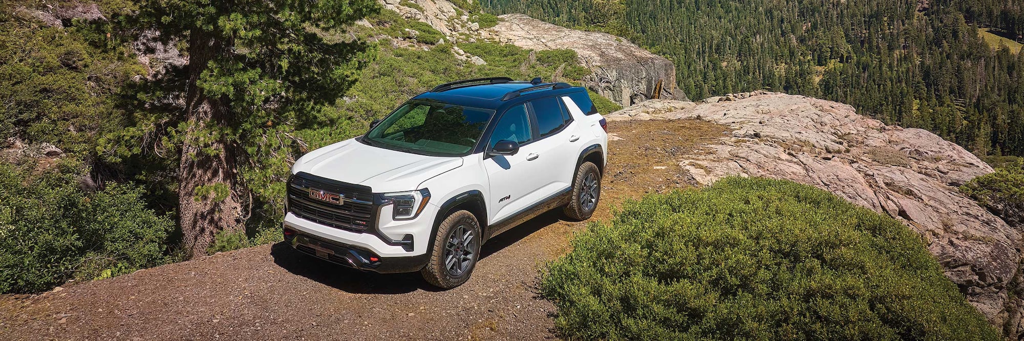 A White GMC Terrain AT4 SUV with a Black Roof Parked on a Dirt Path Overlooking a Forested Valley with Steep Rocky Cliffs