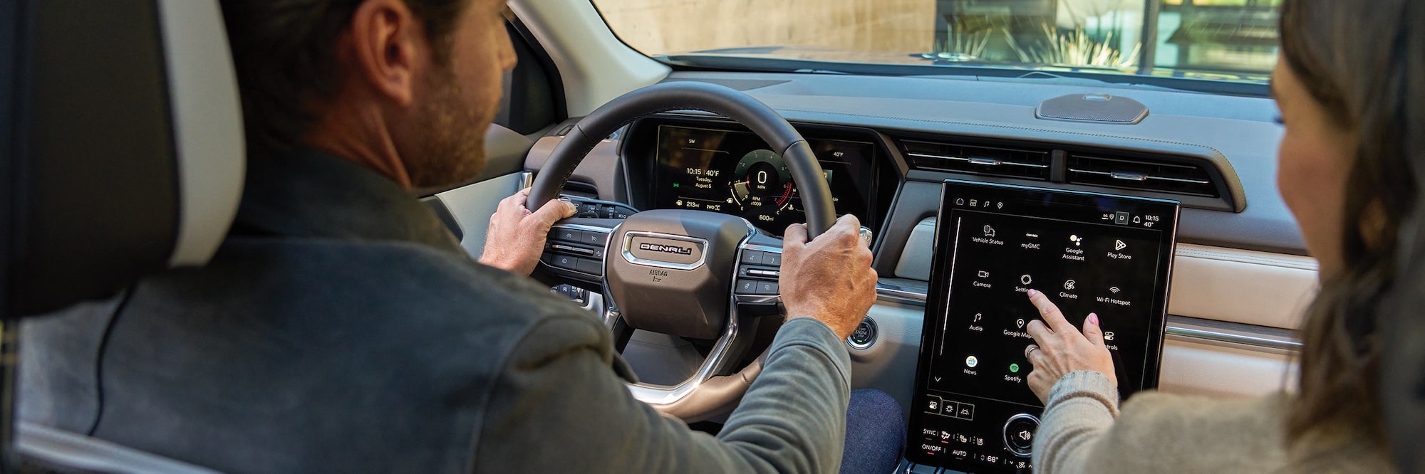 A Man Driving a GMC Denali Vehicle with His Hands on the Steering Wheel while a Woman Uses the Large Touchscreen Infotainment System on the Passenger Side