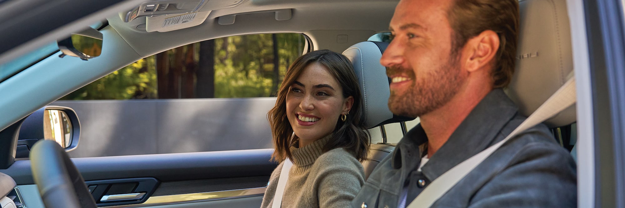 A Smiling Woman with Dark Hair in a Beige Sweater Looks towards a Man Driving Both Inside a Vehicle with Trees Visible Outside the Windows