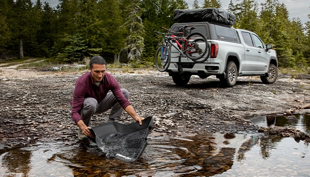 A Man Rinsing his Vehicles Floor Liner in a River with his GMC Vehicle Parked in the Background