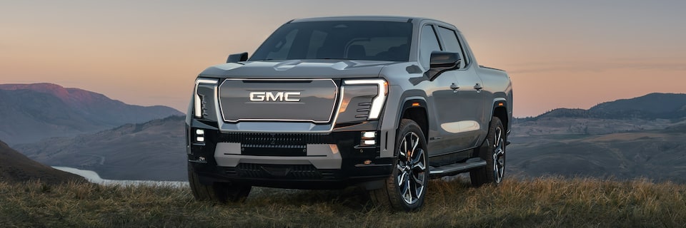 The GMC Sierra EV pickup truck parked in a green valley at sunset with a beautiful mountain view in the distance.