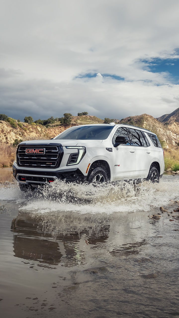 A White GMC Yukon AT4 SUV Drives Through a Shallow Stream, Splashing Water, with Hills and Brush In the Background