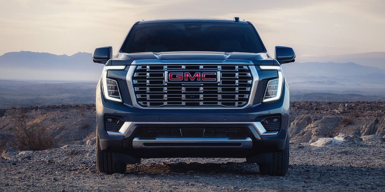 The Front View of a Dark Blue GMC Yukon SUV with a Prominent Grille and GMC Logo, Parked On a Dirt Path At Sunset