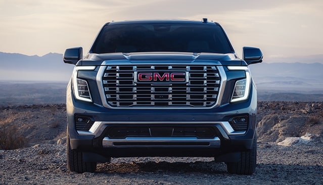 The Front View of a Dark Blue GMC Yukon SUV with a Prominent Grille and GMC Logo, Parked On a Dirt Path At Sunset