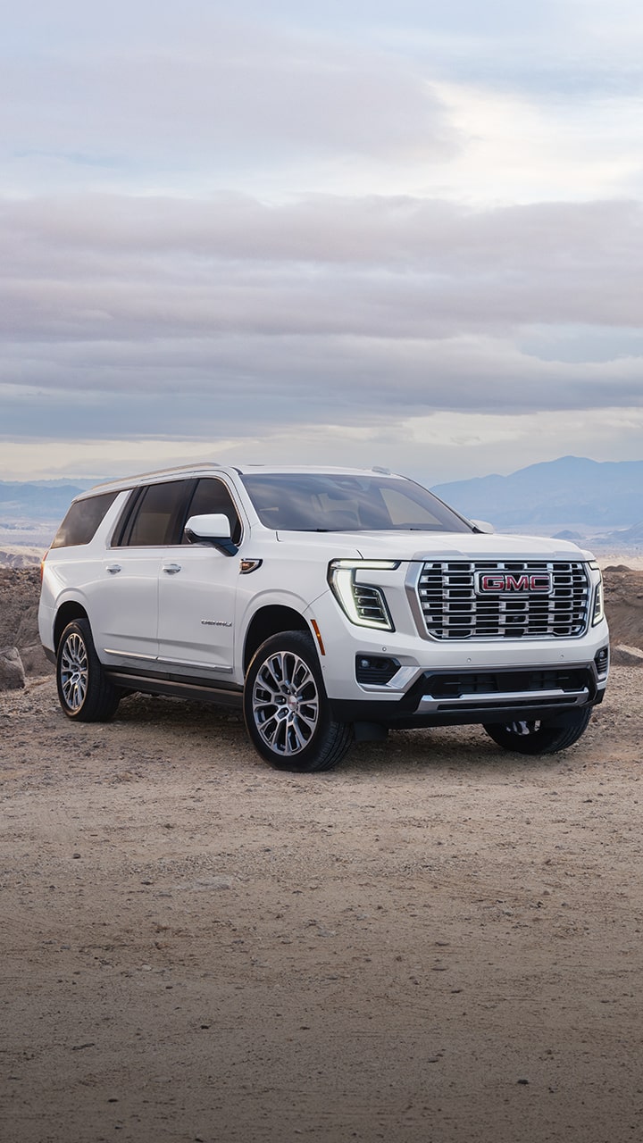 A White GMC Yukon SUV and a Dark Blue GMC Yukon SUV Are Parked Side-by-Side On a Dirt Road Against a Distant Mountain Range